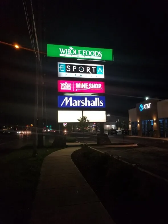 A large pylon sign with multiple light boxes adverting stores in a plaza at night.